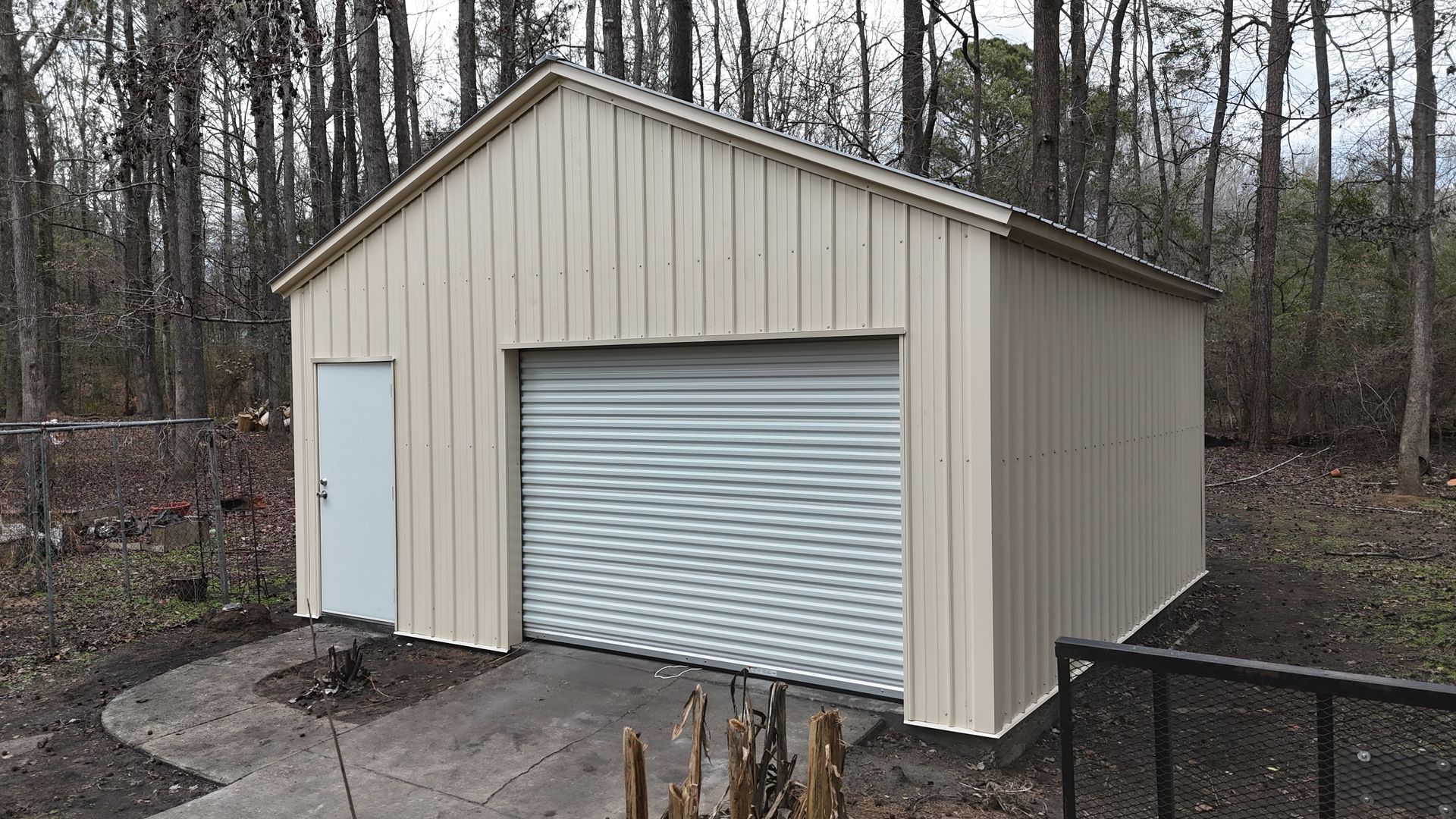 Tan metal garage with a white door and a roll-up garage door in a wooded area.
