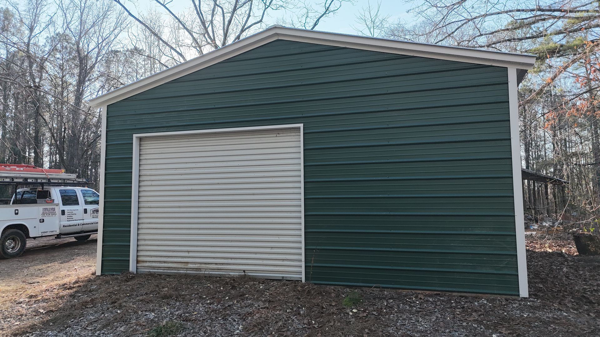 Green metal garage with white garage door, next to a white service truck in a wooded area.