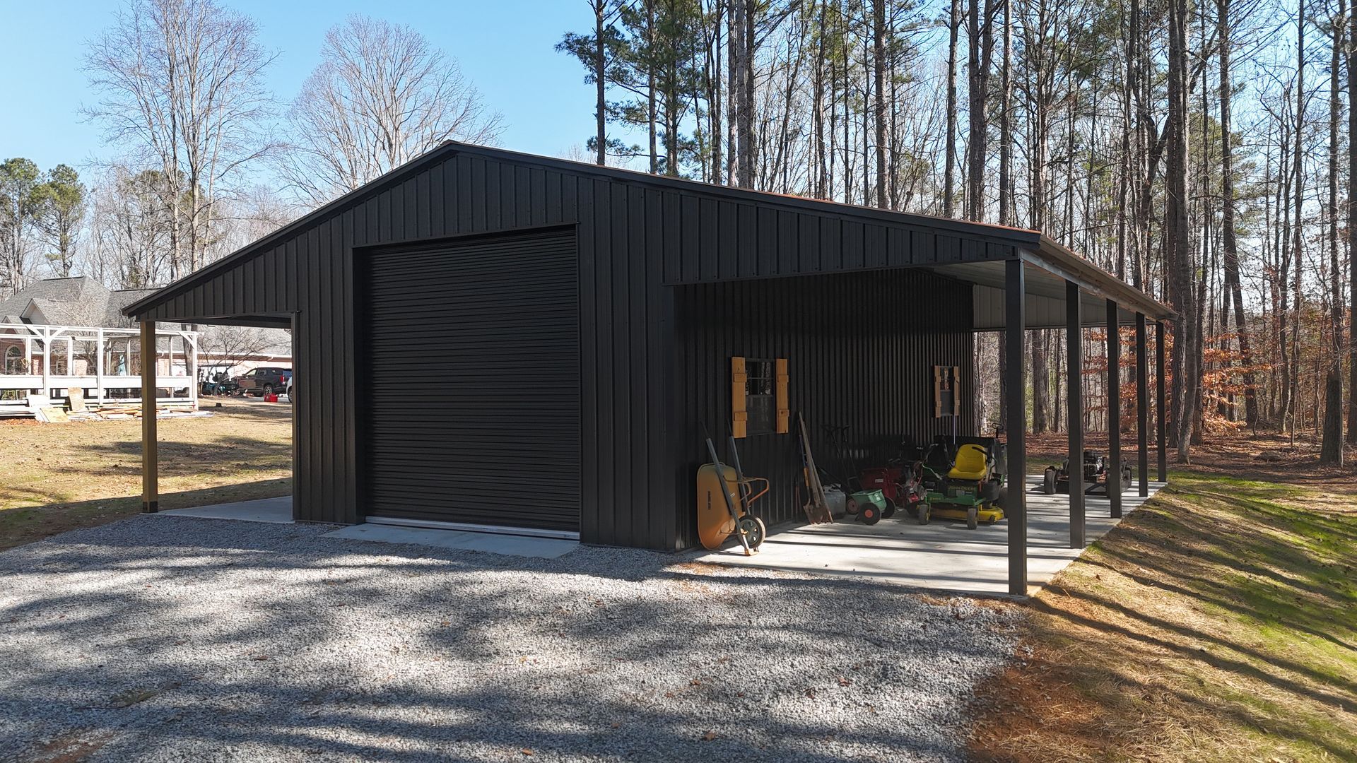 Black metal garage with a carport on a gravel driveway, set in a wooded area.