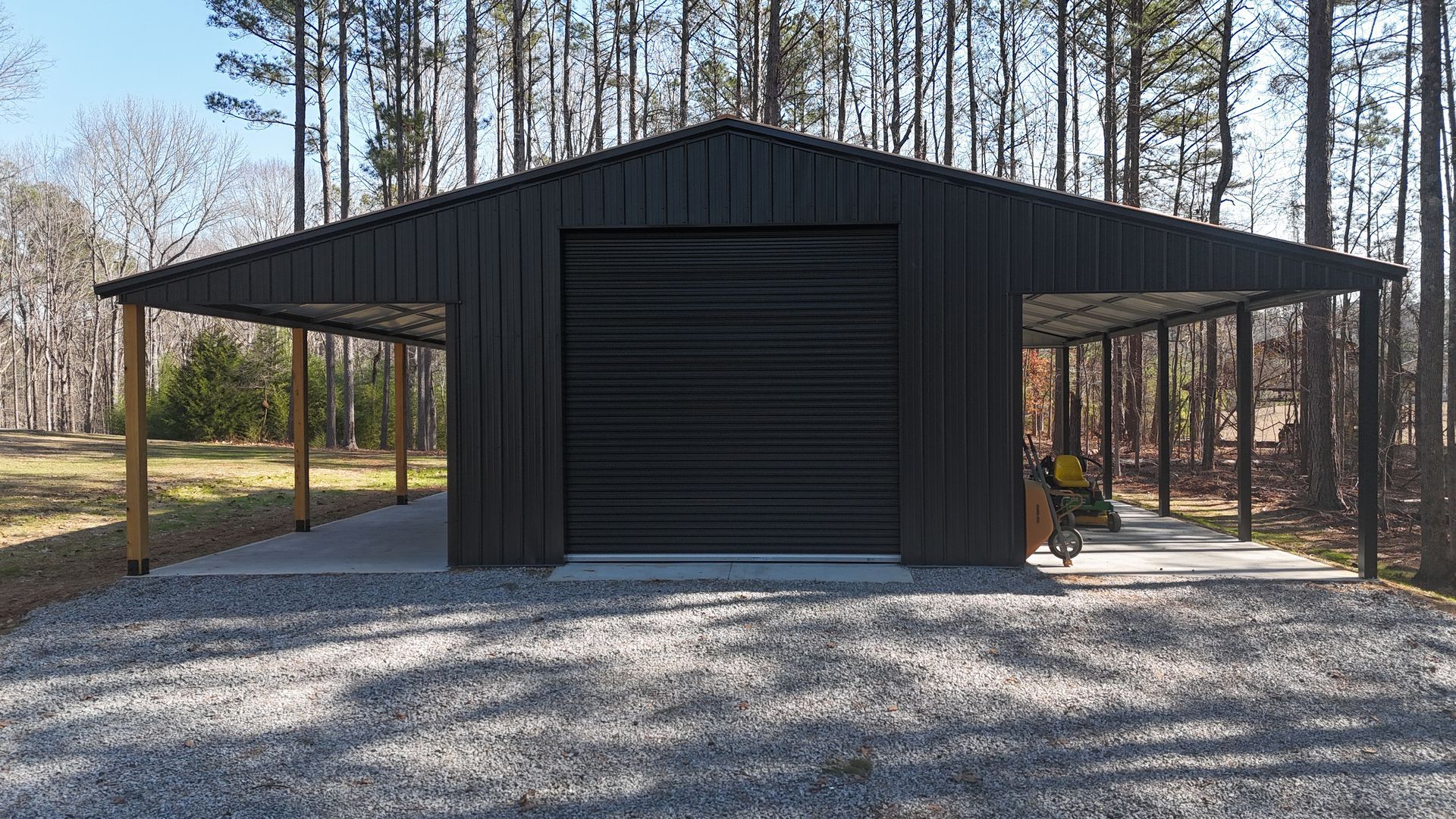 Dark gray metal garage with two side carports on a gravel driveway.