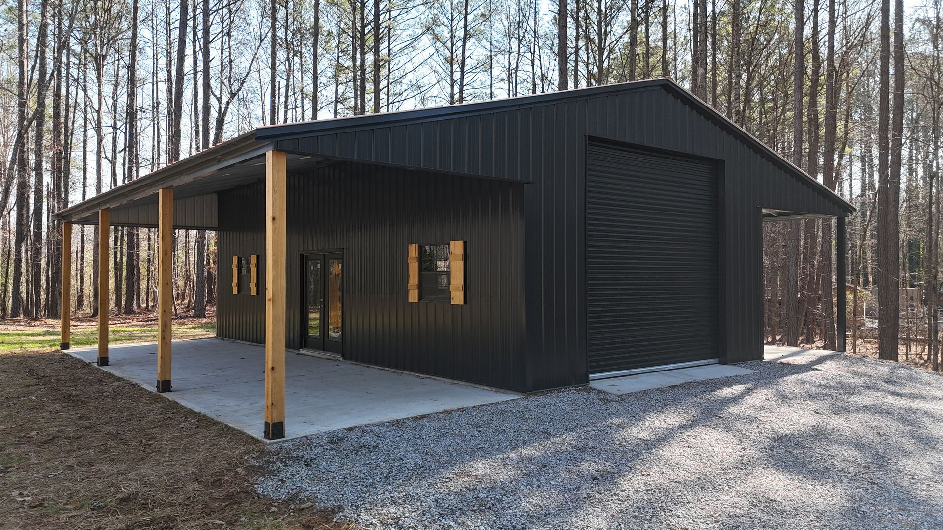 Black metal shed with a covered porch on a concrete slab in a wooded area.