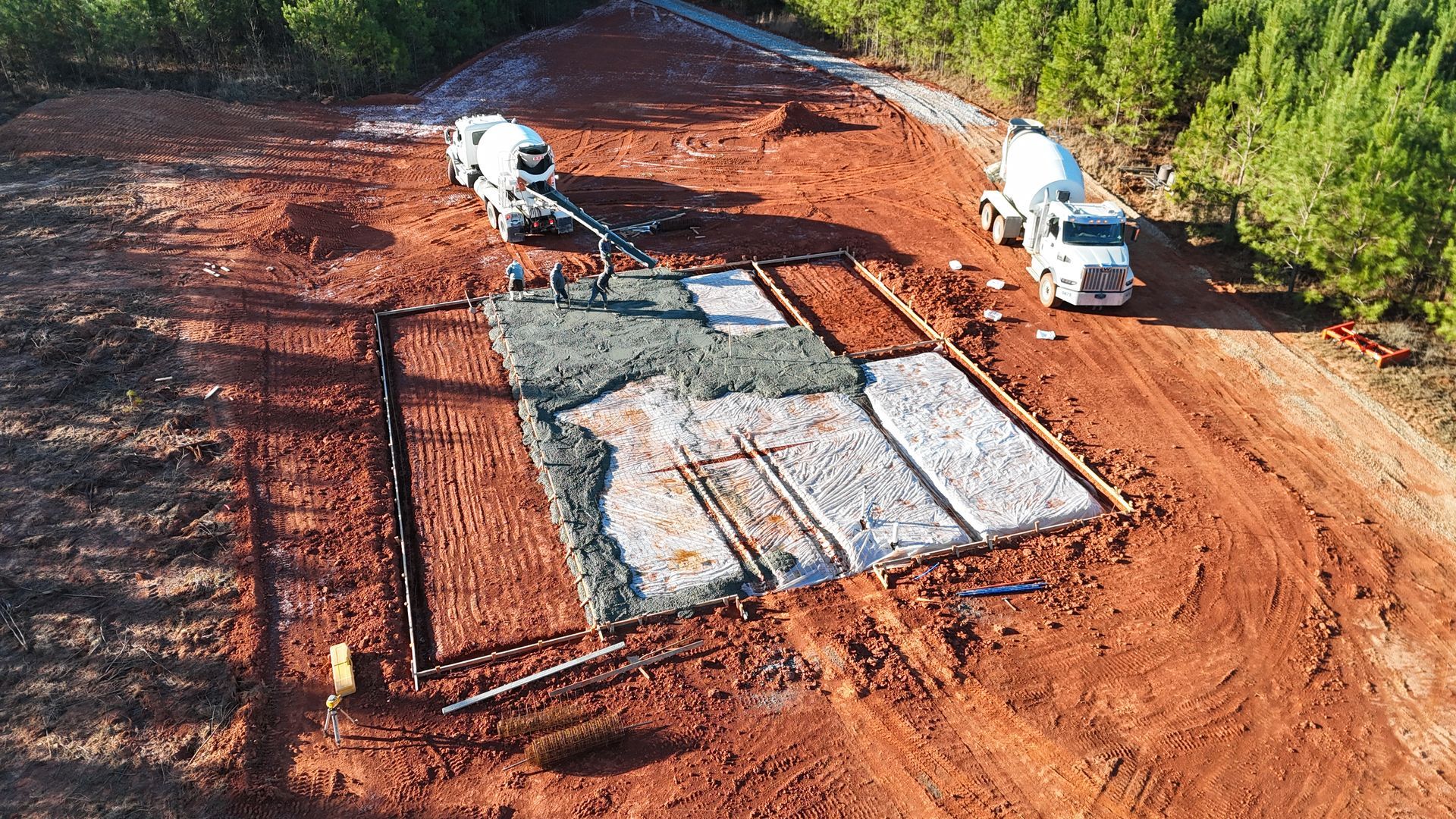 Construction site: concrete pouring for a foundation. Two cement trucks pour into a wooden-framed area on reddish soil.