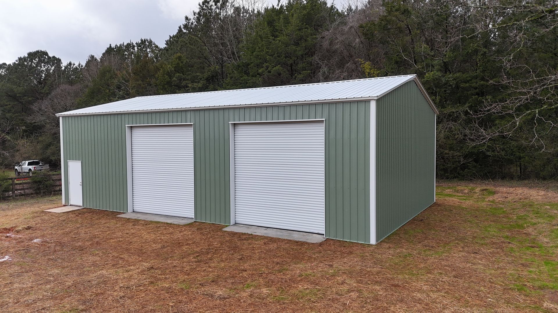 Green metal garage with two doors and a white door, set in a wooded area.