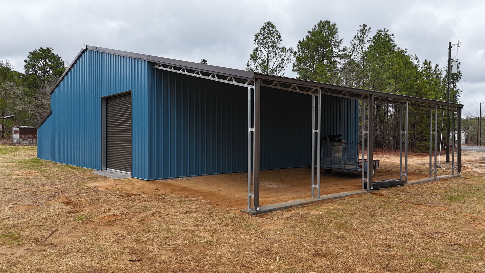Blue metal shed with a covered carport. Located outdoors, in front of a tree line, and on a dirt lot.