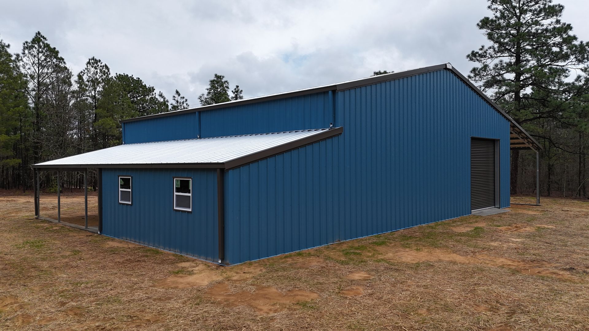 Blue metal barn with a porch and windows, set in a wooded area. Overcast sky.