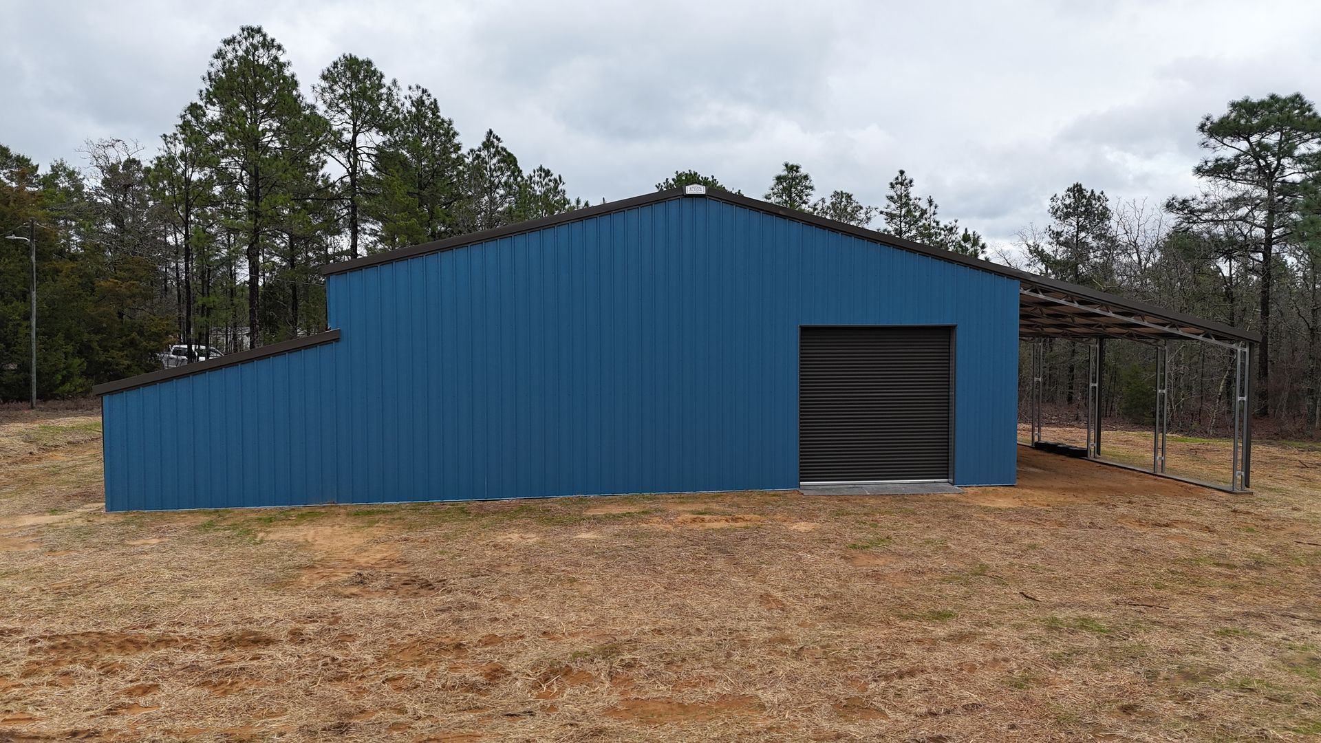 Blue metal barn with a black door and attached carport, in a field with trees.