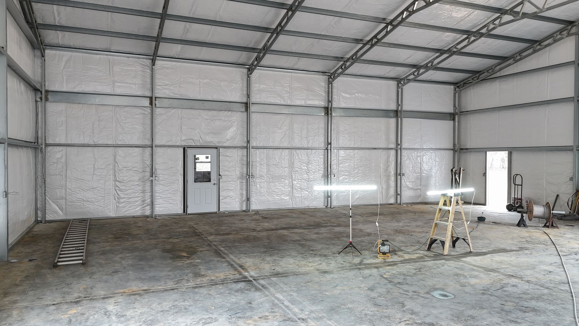 Inside of a large, unfinished metal building. Walls are insulated; concrete floor. Door and light visible.