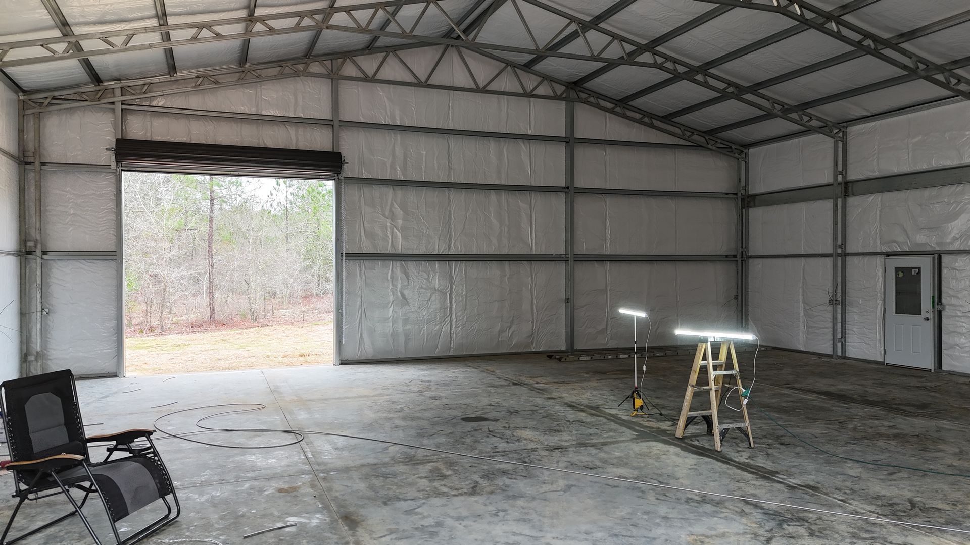 Interior of a metal building with concrete floor and roll-up door. A chair and work lights are visible.