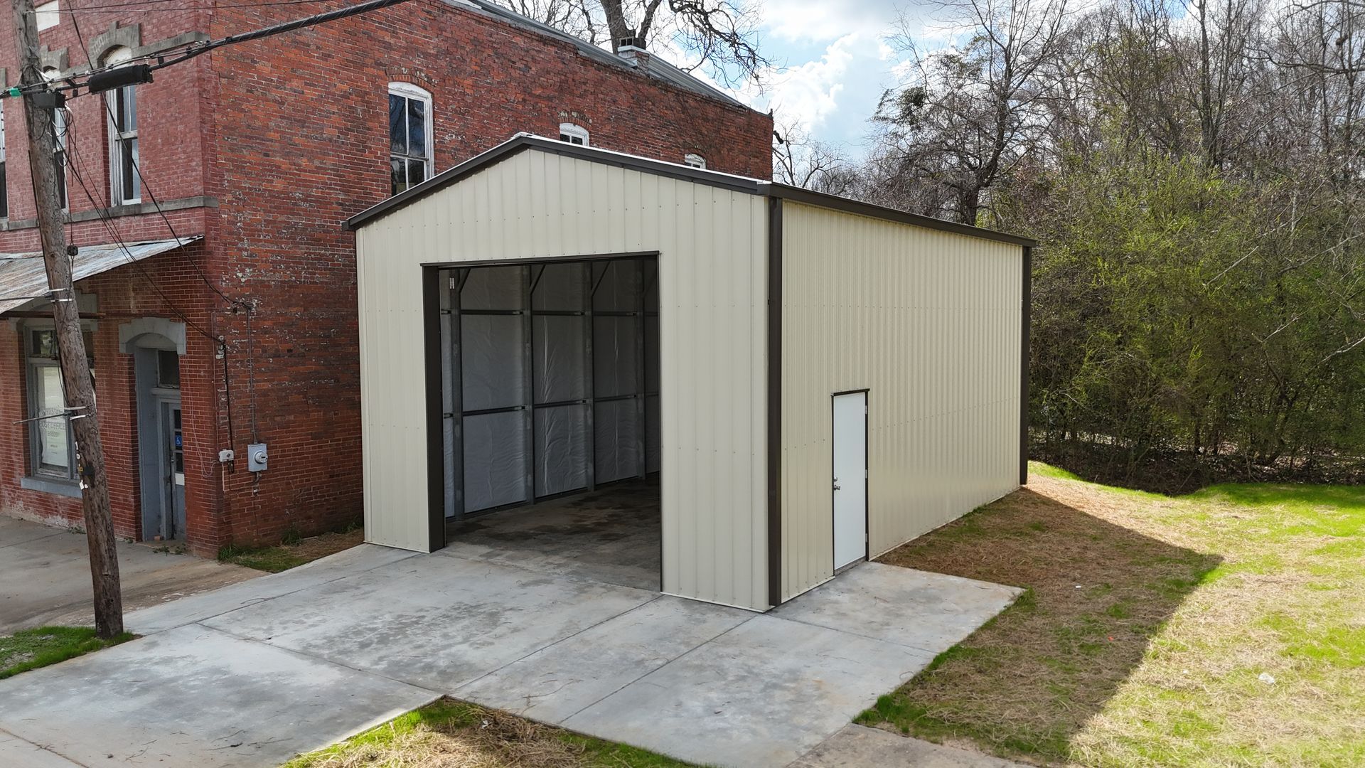A tan metal garage with a side door, built on a concrete slab next to a multi-story brick building.