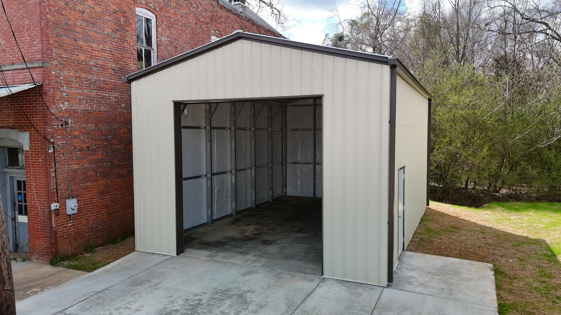 An open beige metal garage structure stands next to a weathered red brick building on a concrete driveway.