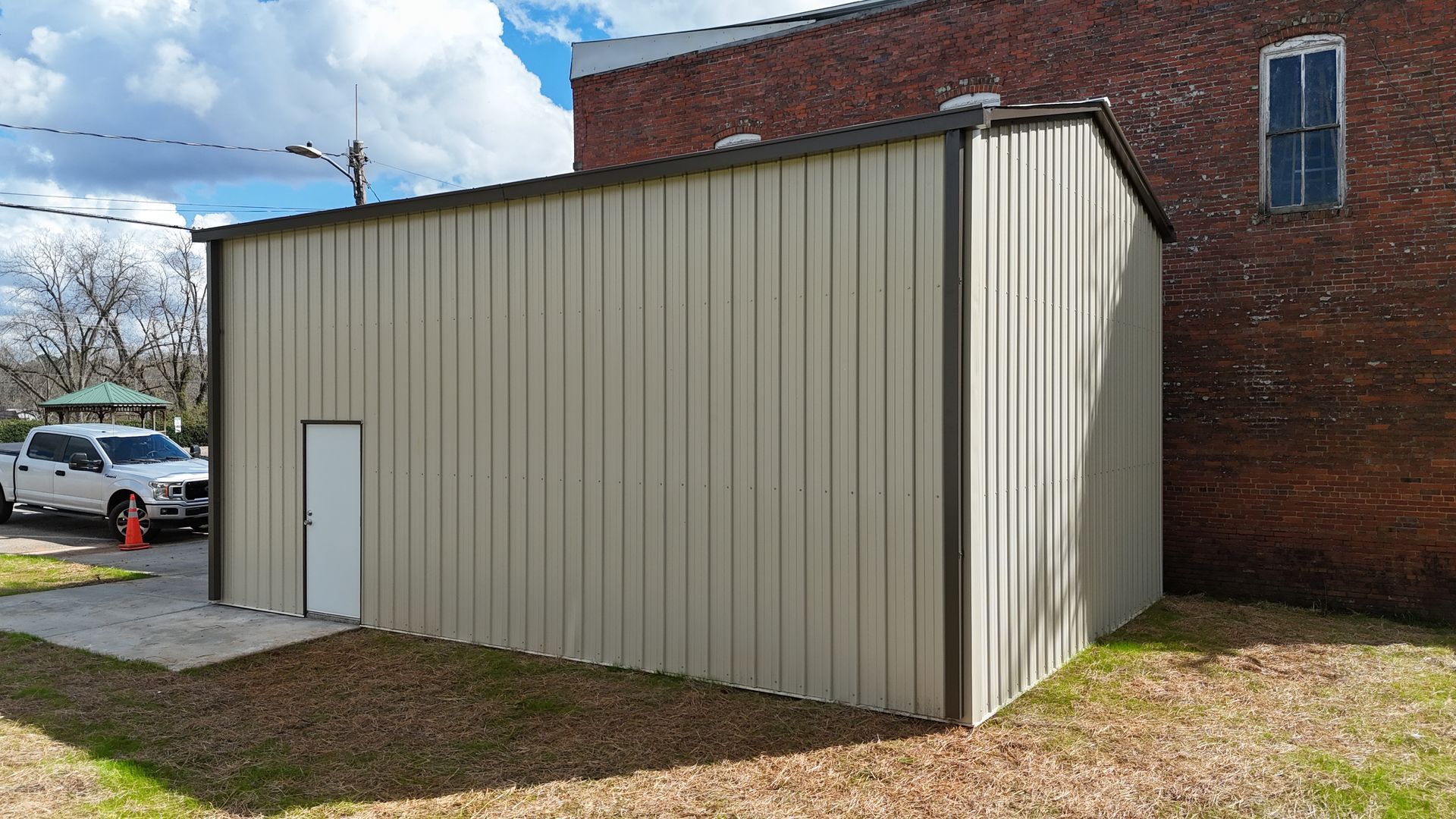 A tan metal storage shed stands next to a red brick building outdoors with a white truck parked in the background.