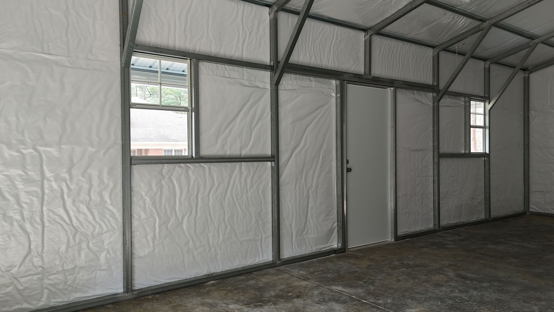 The interior of a metal building with white insulation, two windows, and a white walk-through door on a concrete floor.