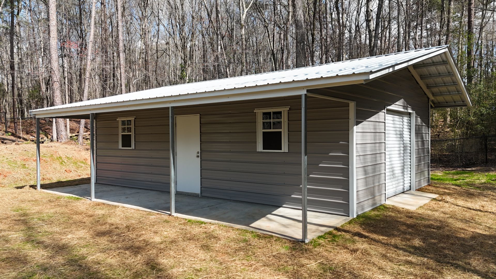 Gray metal workshop building with a white door, two windows, and a front porch, situated in a wooded area.