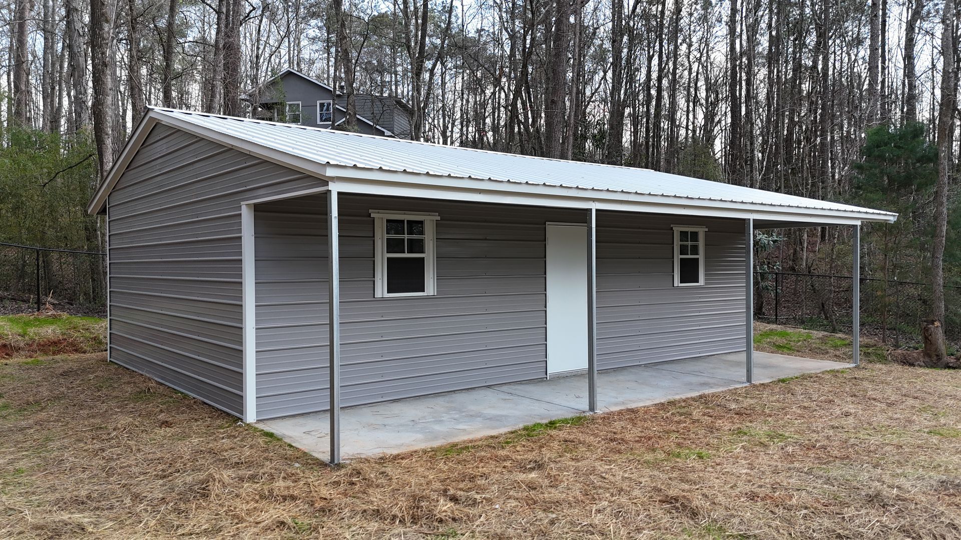A gray metal storage building with a front porch, white trim, two windows, and a door, set in a wooded area.