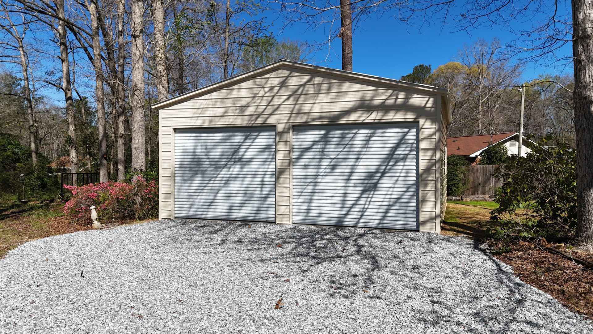 A beige metal two-car garage with white roll-up doors, set on a gravel driveway surrounded by trees under a blue sky.
