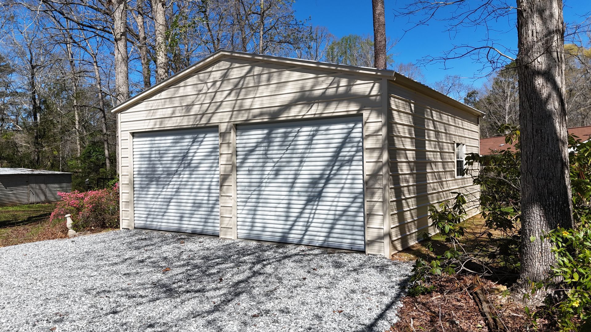 A beige metal two-car garage with white roll-up doors, set on a gravel driveway surrounded by trees.