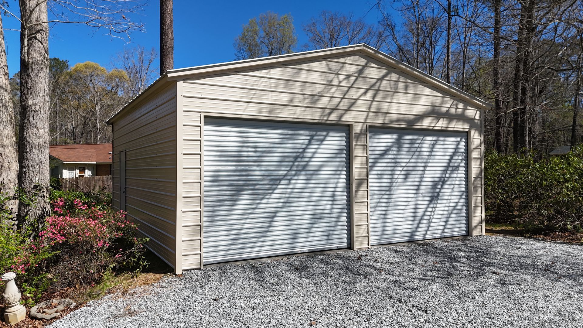 A beige metal two-car garage with white roll-up doors sitting on a gravel driveway in a wooded area.