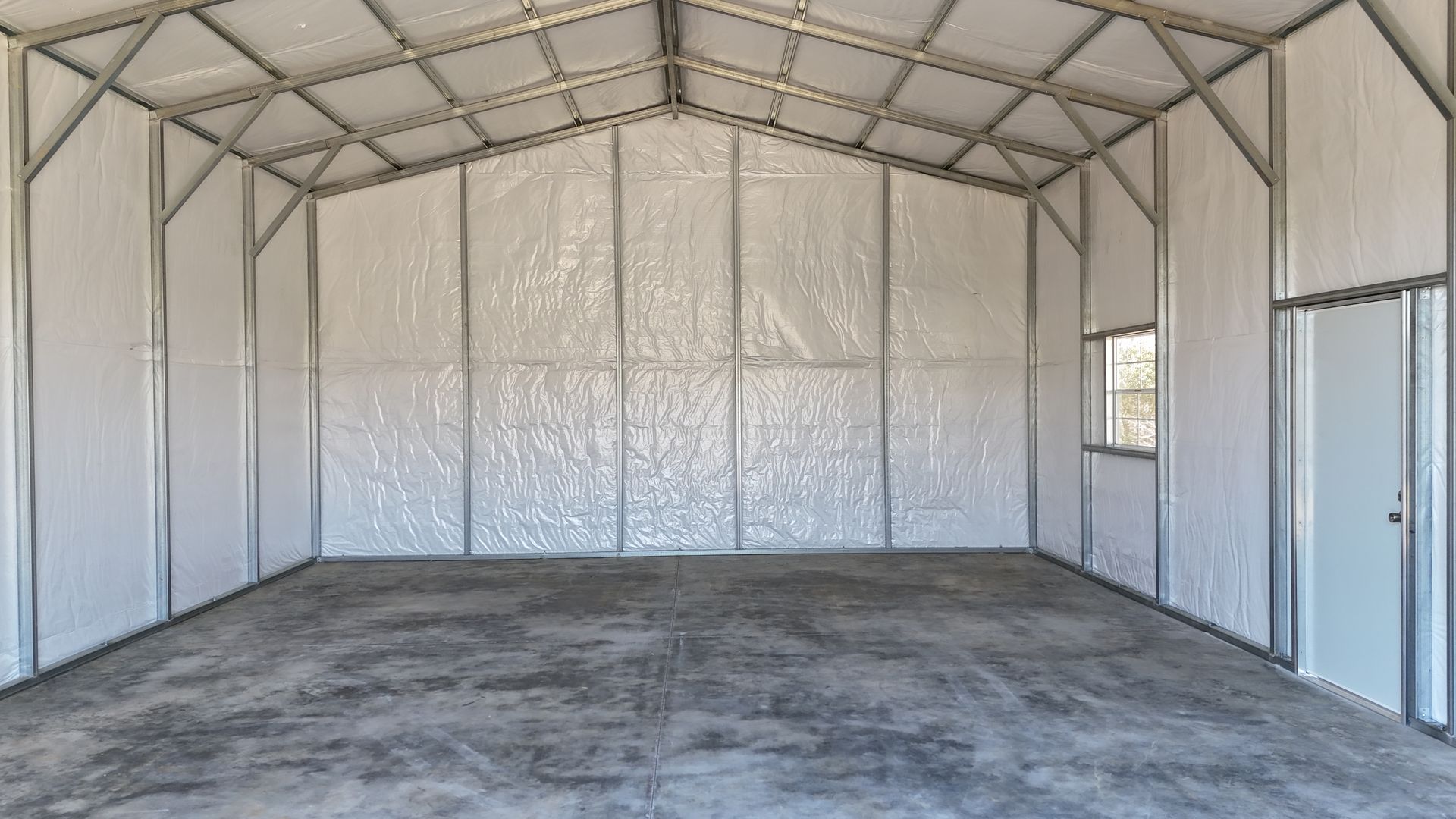 Interior of a wide, empty metal building with a concrete floor, white insulated walls, and a side door and window.