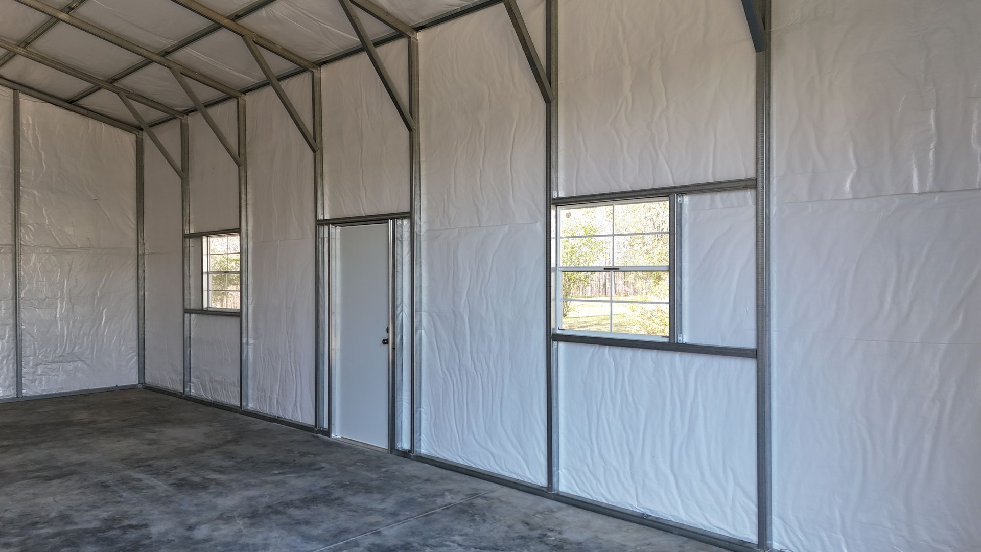 The interior of a metal building with concrete floors, white-insulated walls, a side door, and two rectangular windows.