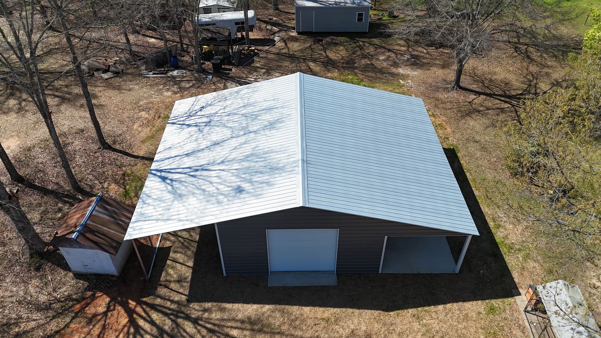Aerial view of a gray metal garage with a white roof and extended awning, set in a wooded lot with another small shed.