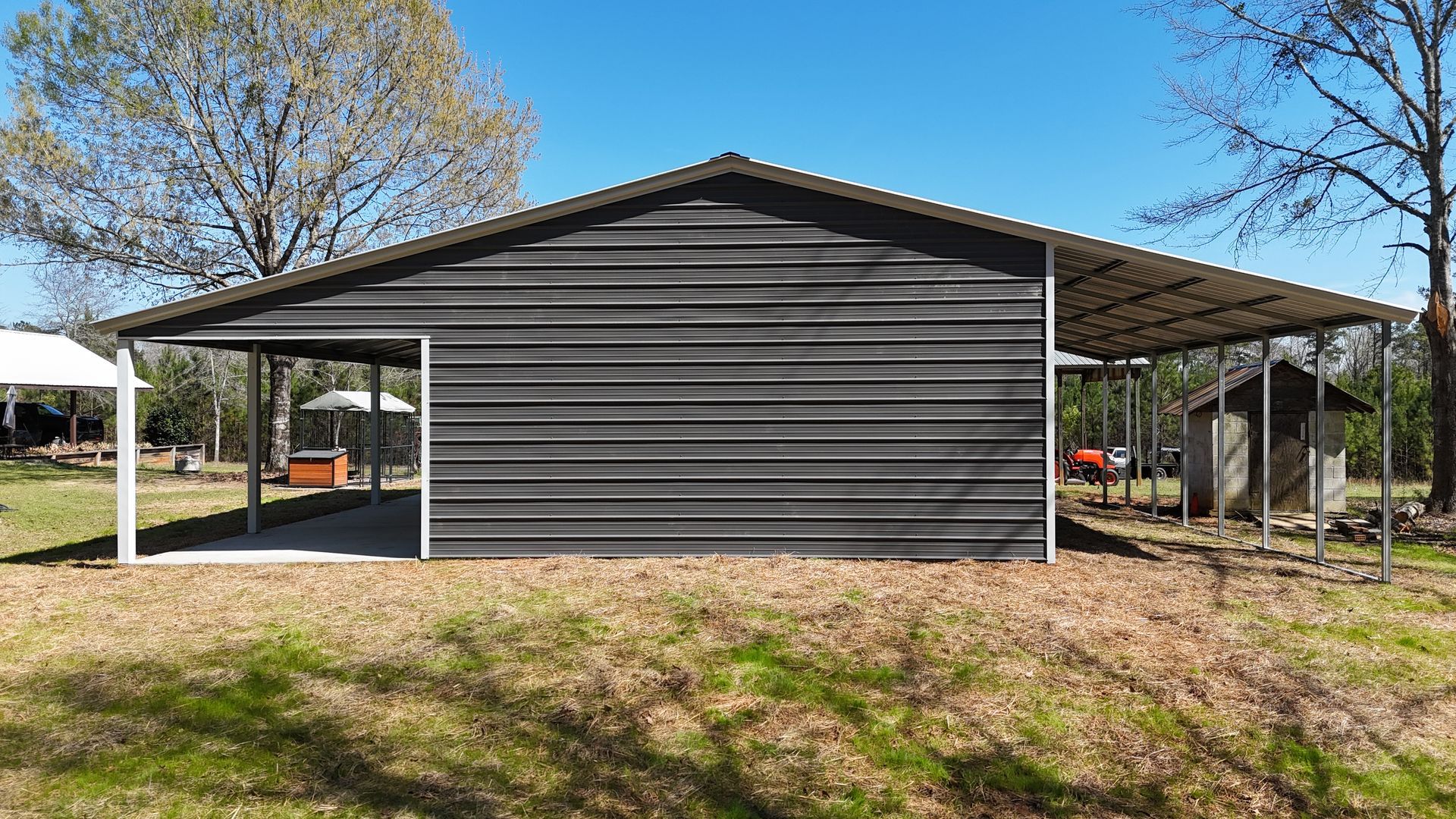 A dark gray metal barn with an open-sided lean-to on both sides, situated in a grassy yard under a clear blue sky.