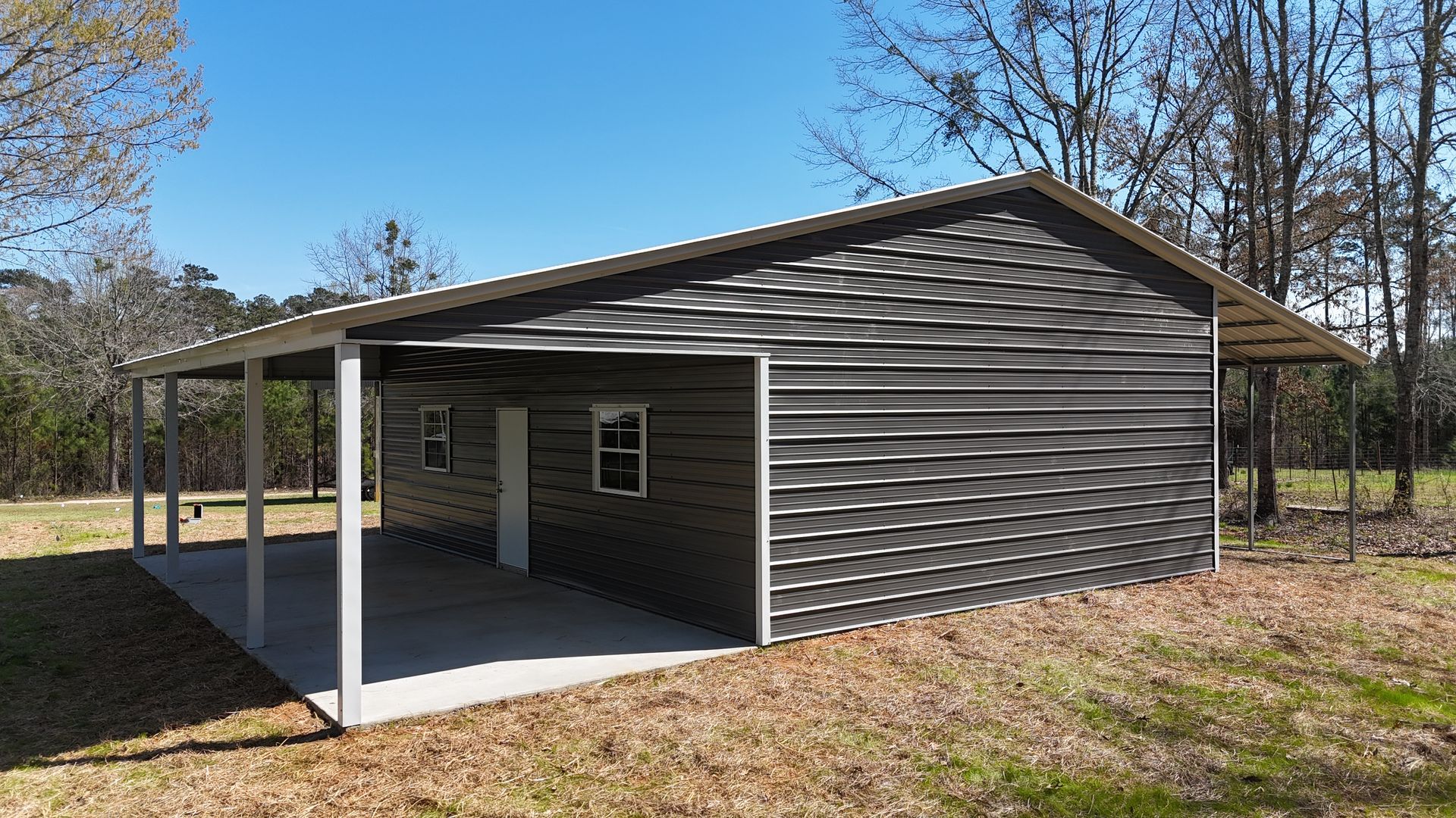 A gray, modern metal building with a side porch, windows, and a doorway, situated on a grassy lot with trees nearby.