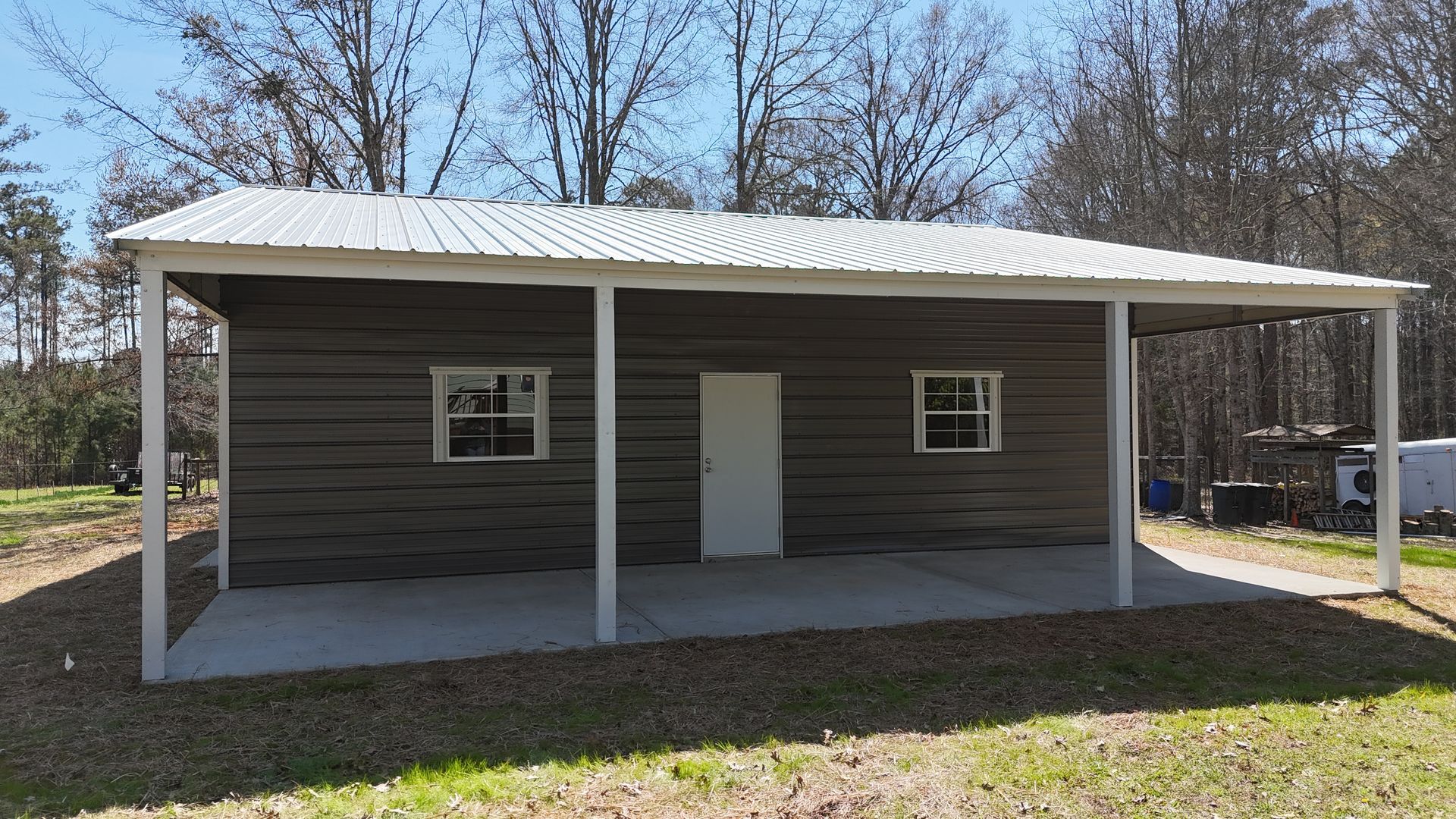 A rectangular shed with horizontal dark brown siding, a white metal roof, a white door, two windows, and a front porch.