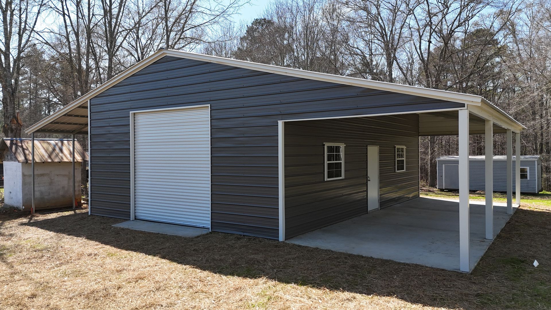 Dark gray metal workshop building with a roll-up door, side porch, and concrete slab, set in a wooded rural area.