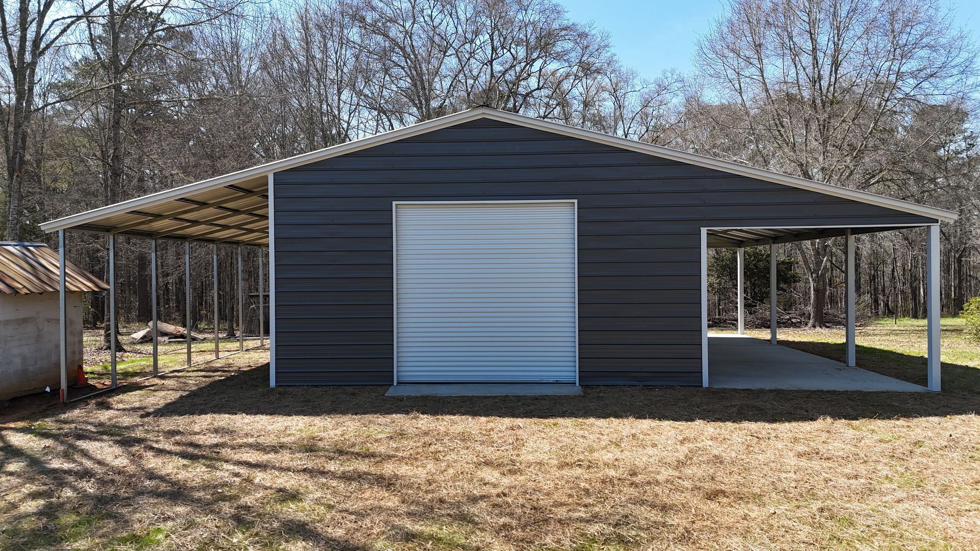 A dark gray metal garage with a white roll-up door and open carports on both sides, situated in a wooded yard.