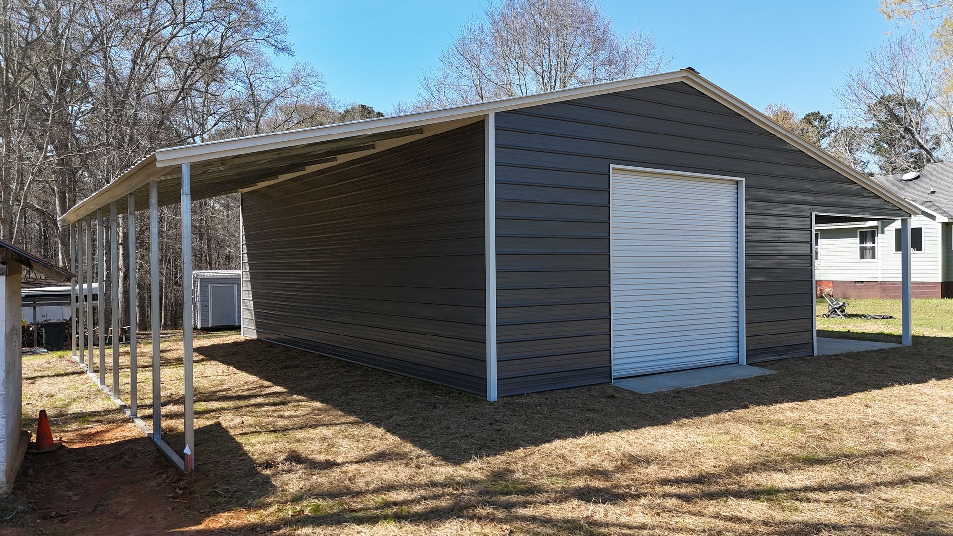 A new dark gray metal garage building with a side carport on a grassy, partially wooded lot.