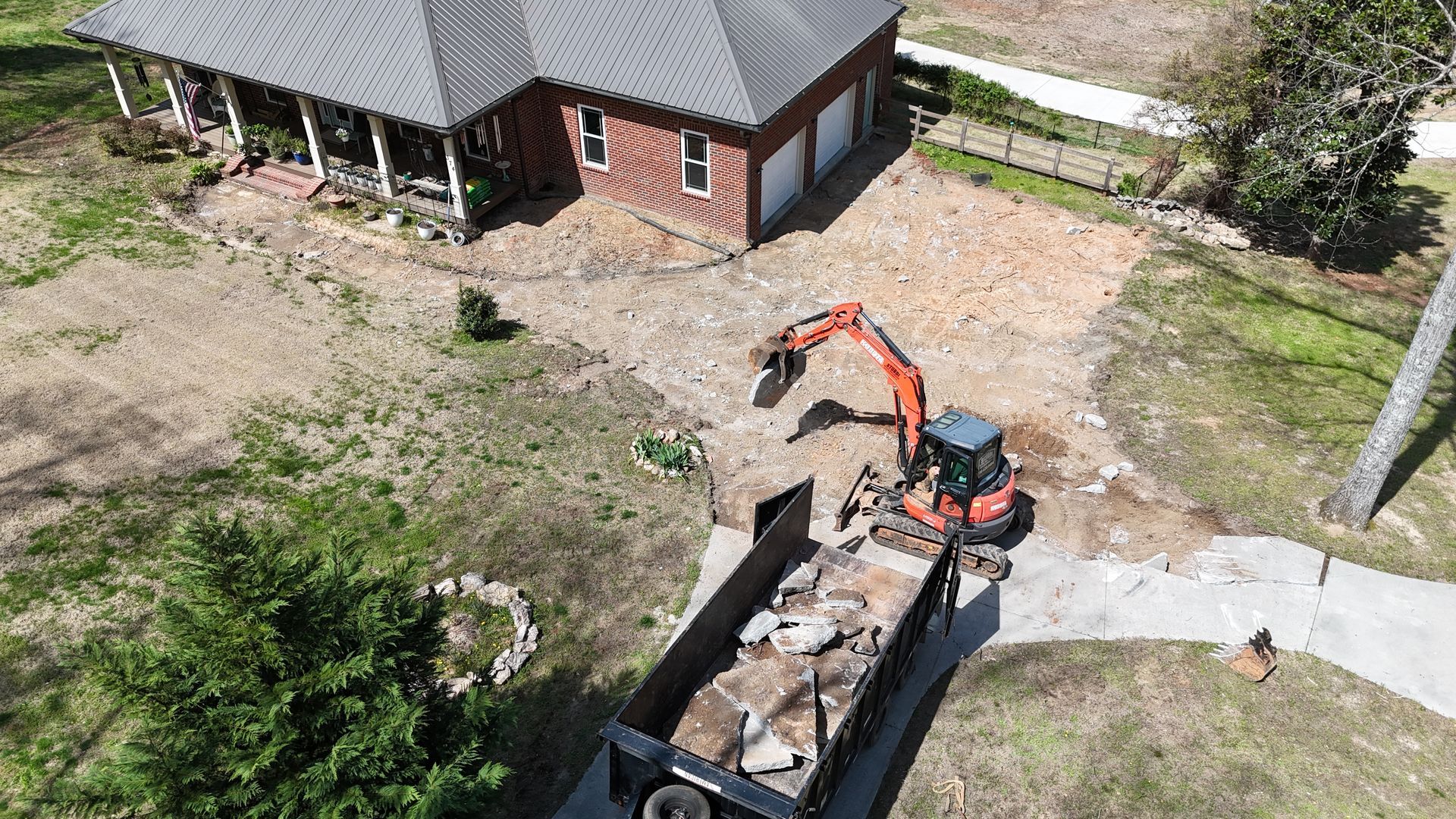 An orange excavator loads debris into a dumpster in front of a brick house during a landscape renovation project.