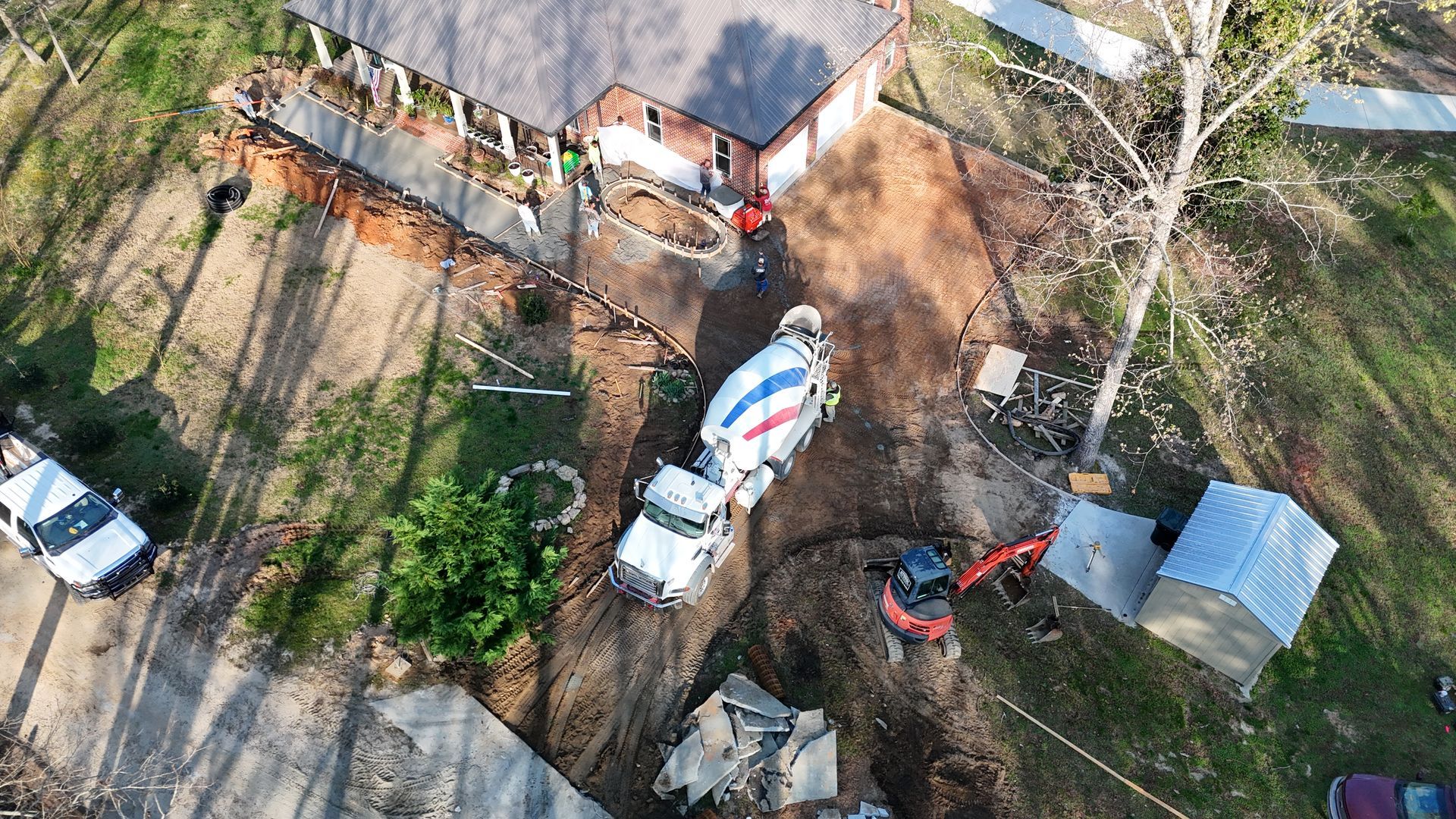 An aerial view shows a concrete truck, a small excavator, and workers near a residential home during construction.