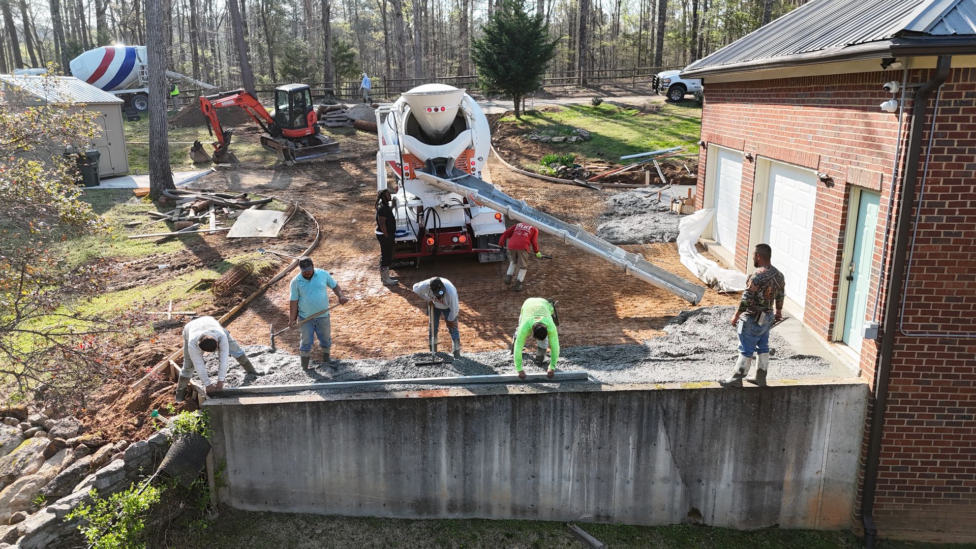 Construction workers pour concrete from a truck mixer into a trench alongside a brick building.