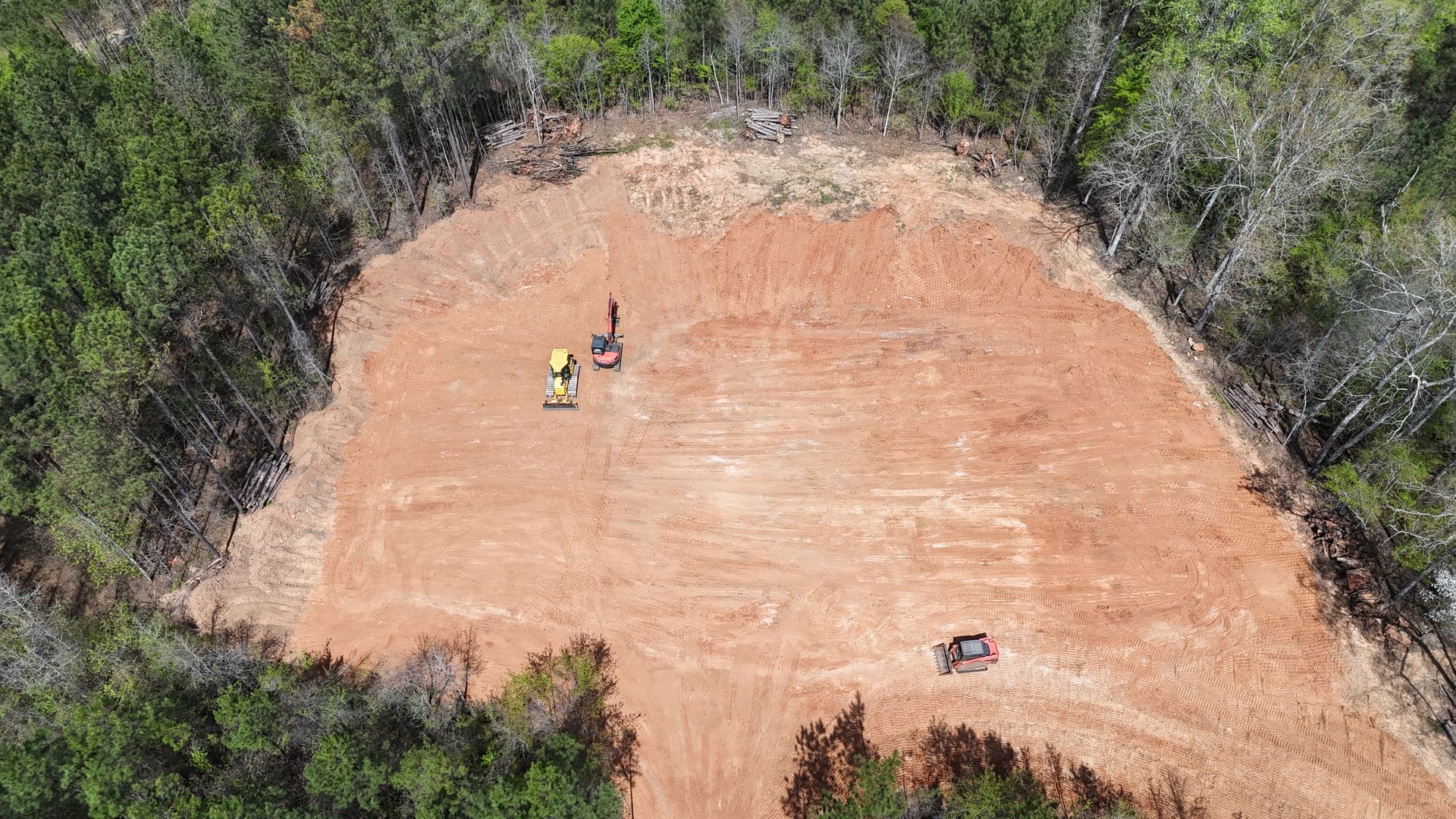 Aerial view of a cleared patch of land surrounded by trees, with two small construction vehicles working on the dirt site.