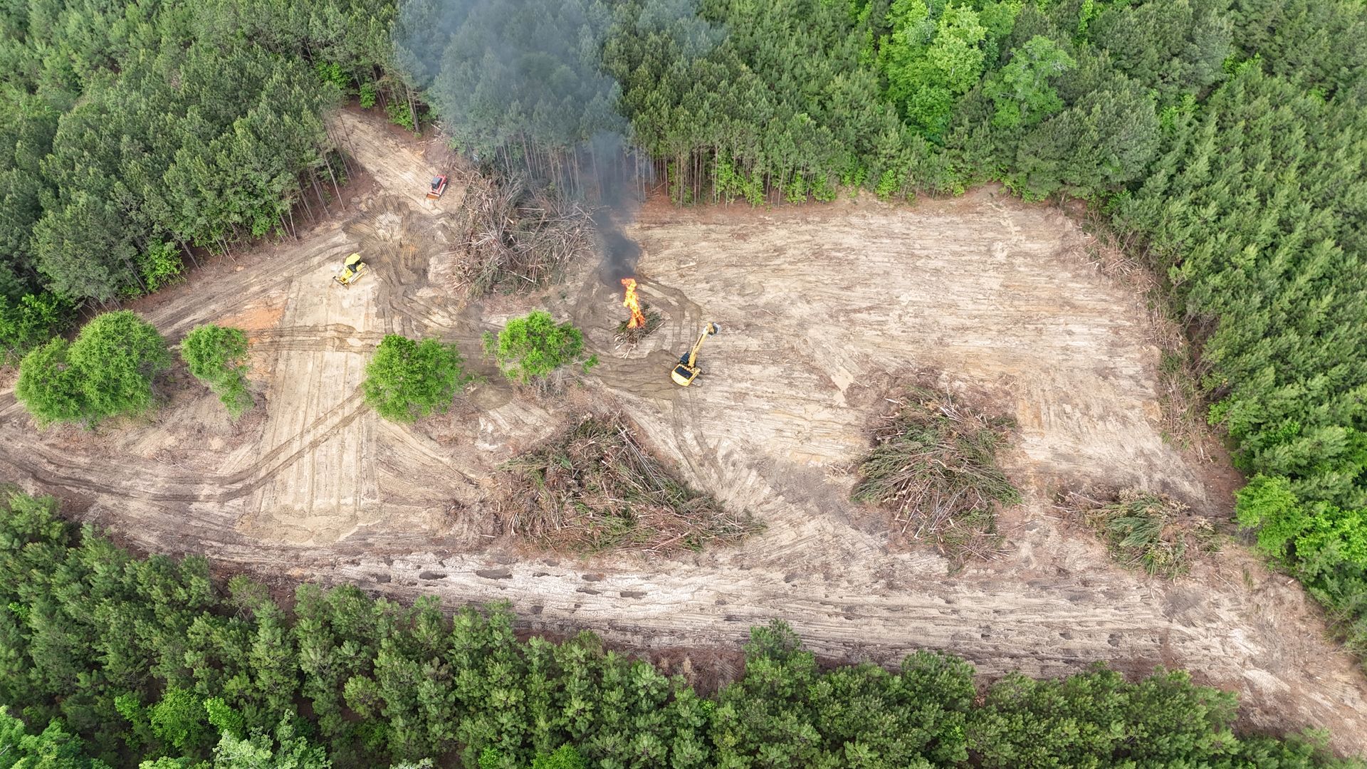 Aerial view of a forest clearing with a small fire and smoke beside exposed dirt and surrounding green trees