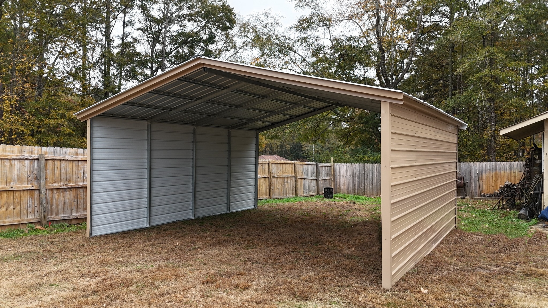 Tan and silver metal carport in a backyard with a wooden fence.