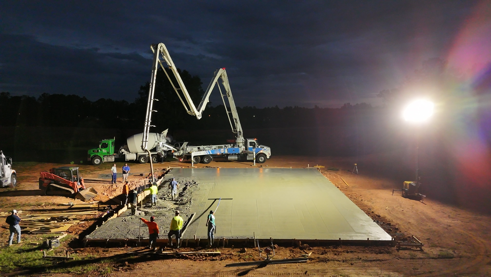 Concrete pour at night with trucks, pump arms, and workers leveling the surface.
