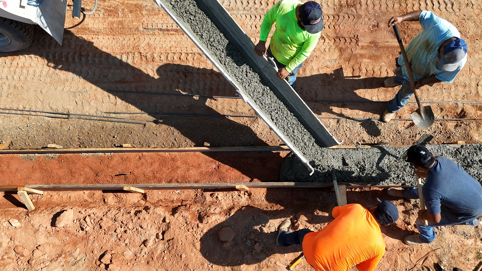 Construction workers pouring concrete into a trench. One worker guides the chute, others shovel and smooth the concrete.