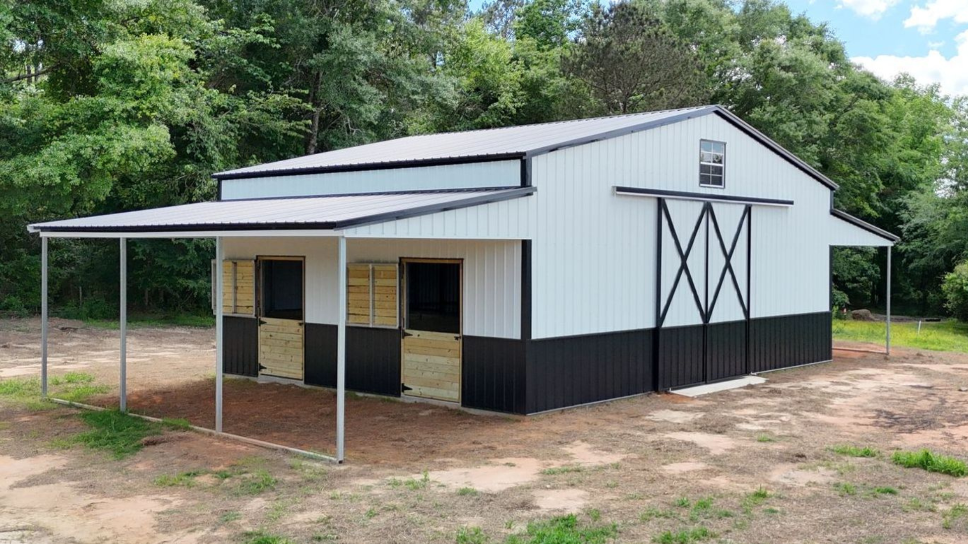 White and black barn with a covered area, in a grassy clearing, surrounded by trees.