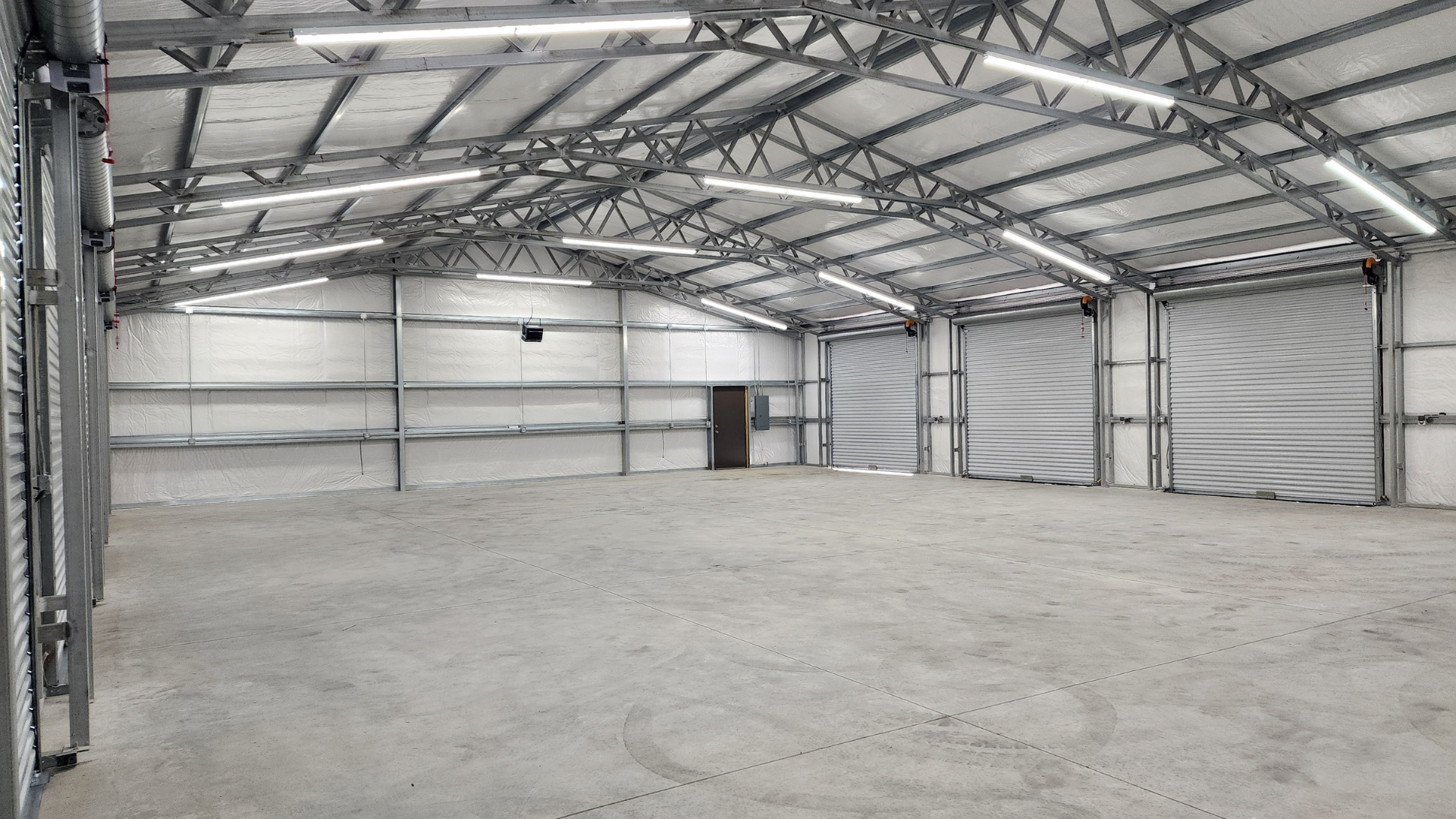 Empty industrial warehouse with metal frame and roll-up doors; concrete floor, bright overhead lighting.