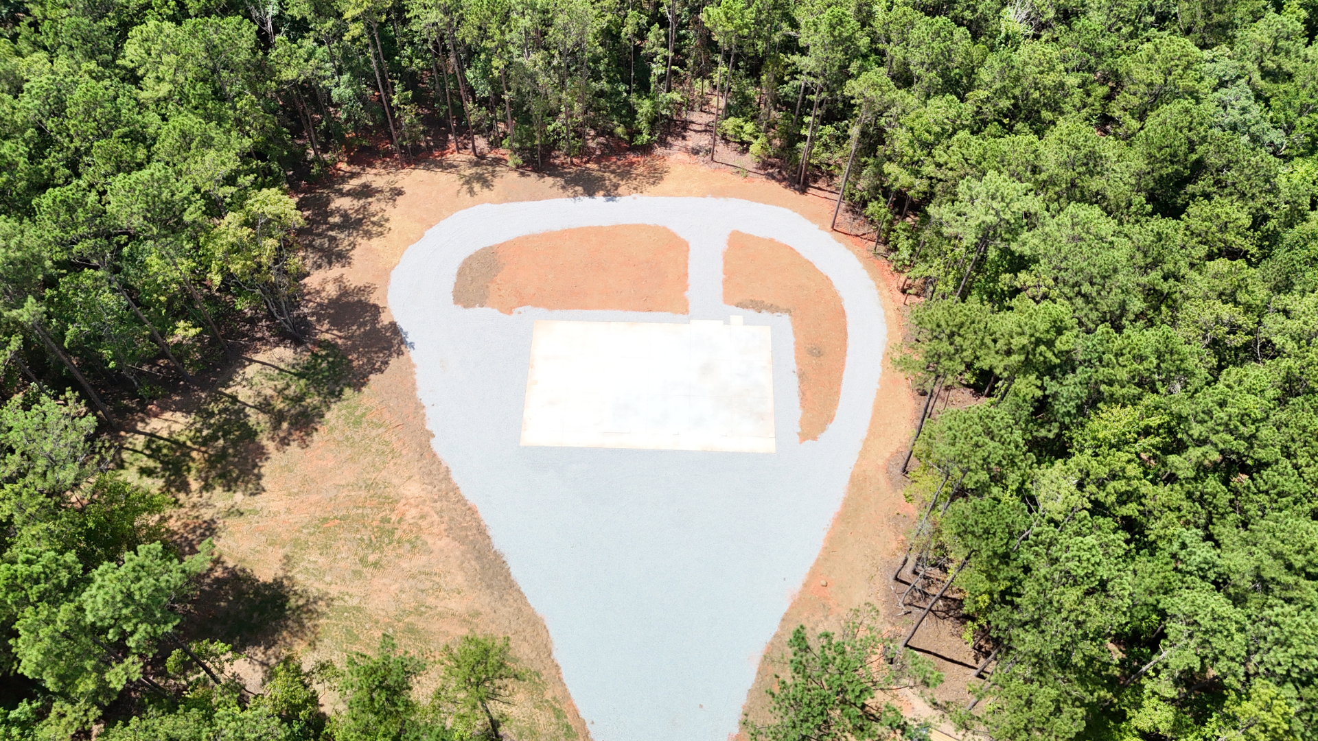 Aerial view: gravel driveway leads to a cleared, rectangular building site surrounded by lush green trees.