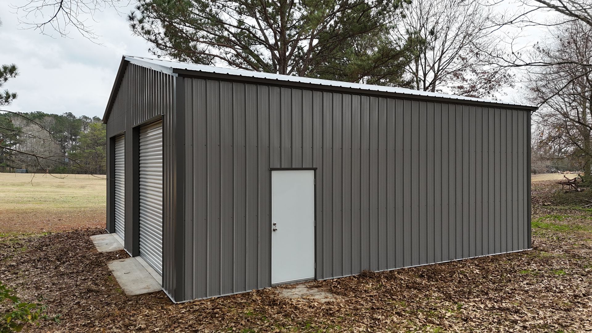 Gray metal garage with two garage doors and a white door, set in a grassy area with trees.