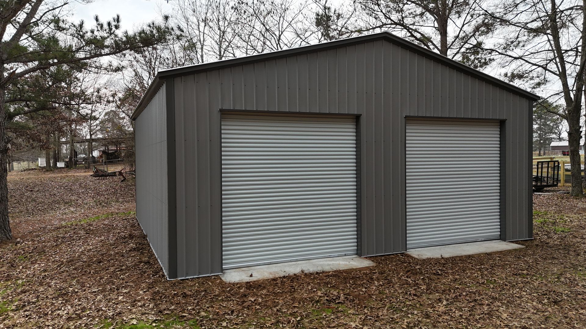 Gray metal two-car garage with roll-up doors, set in a yard with trees and fallen leaves.