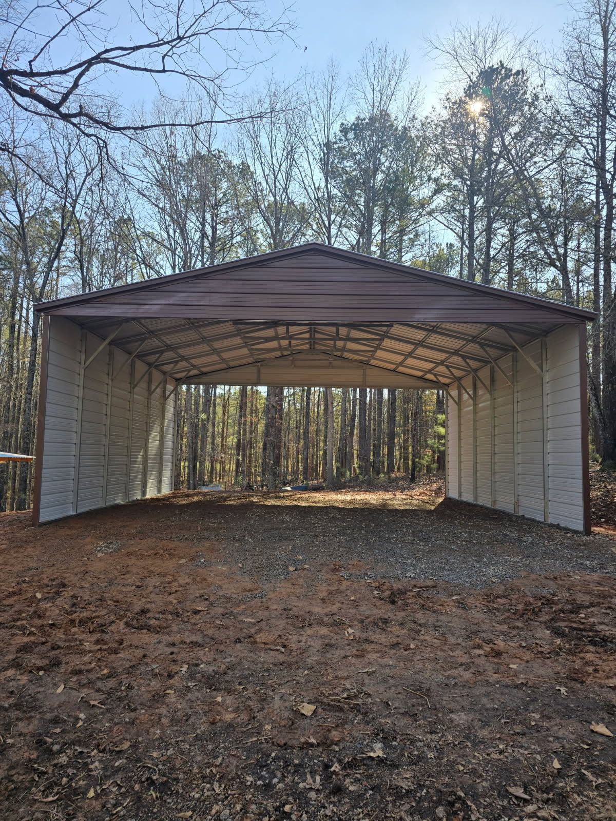 Metal carport with brown roof, beige walls, gravel floor, set in a wooded area, bright sun.