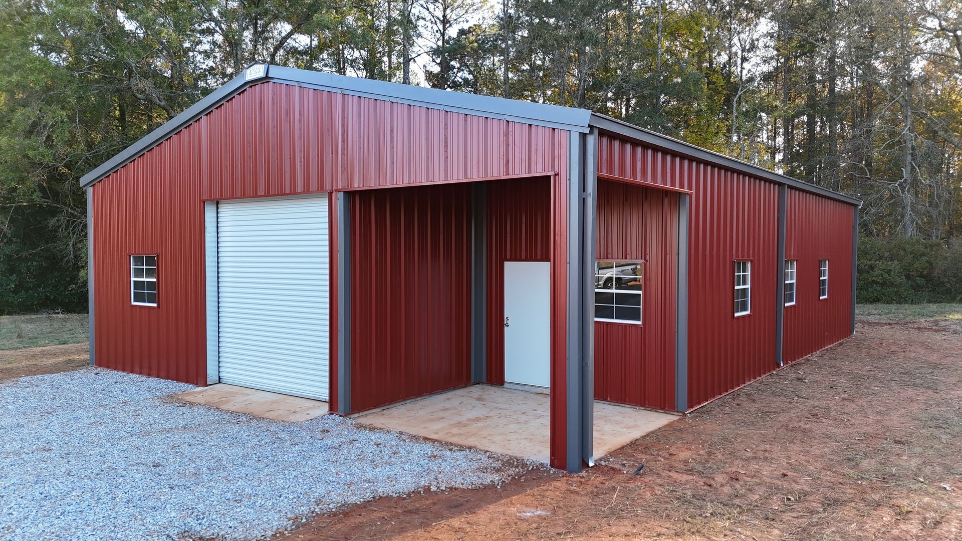 Red metal building with garage door, awning, and windows on a gravel lot.