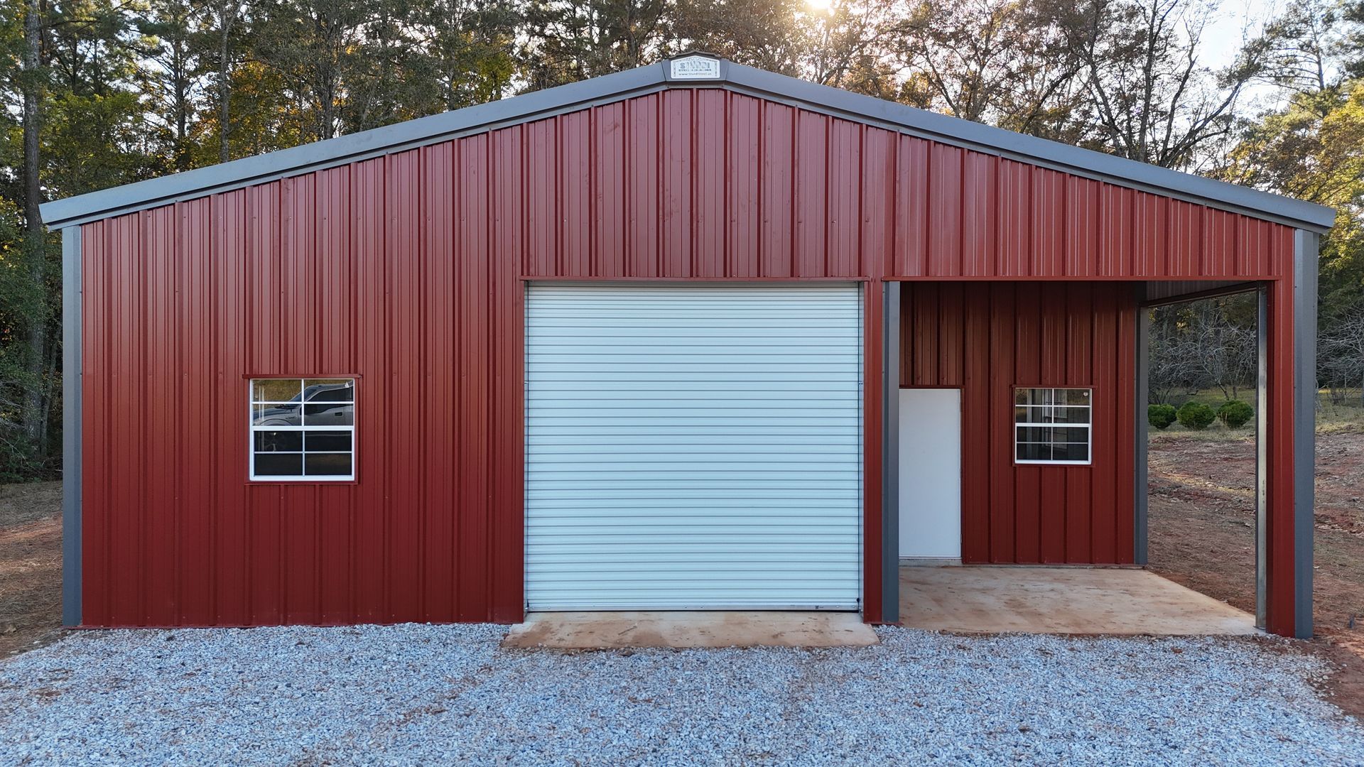 Red metal garage with a light blue garage door and carport, set on gravel.