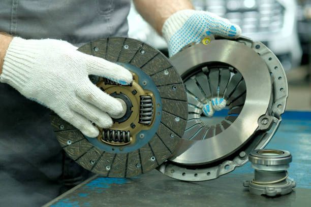 Hands in gloves holding a clutch disc and pressure plate, parts of a car's manual transmission.