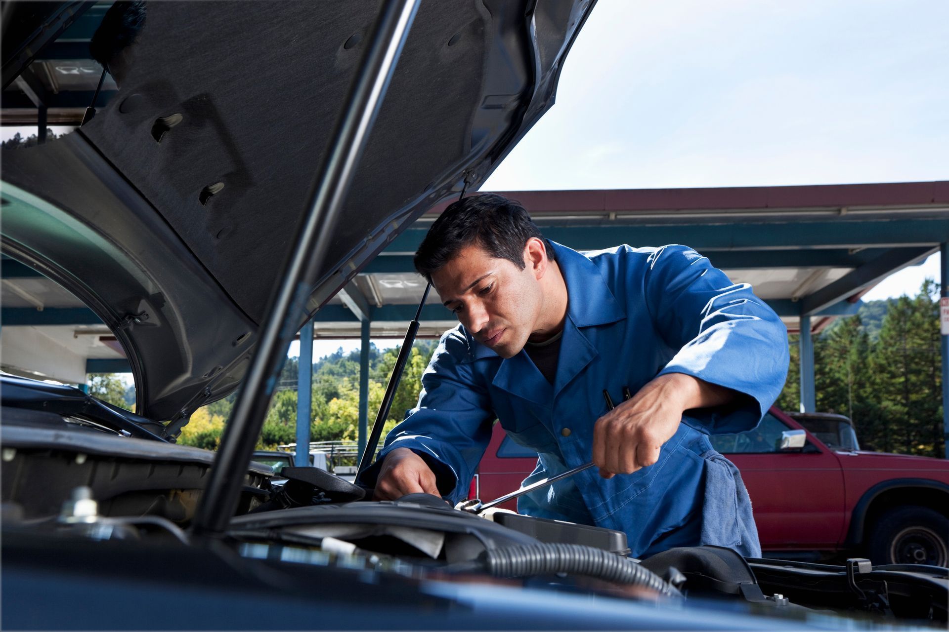 Mechanic in blue jumpsuit works on car engine outdoors, sunny day.