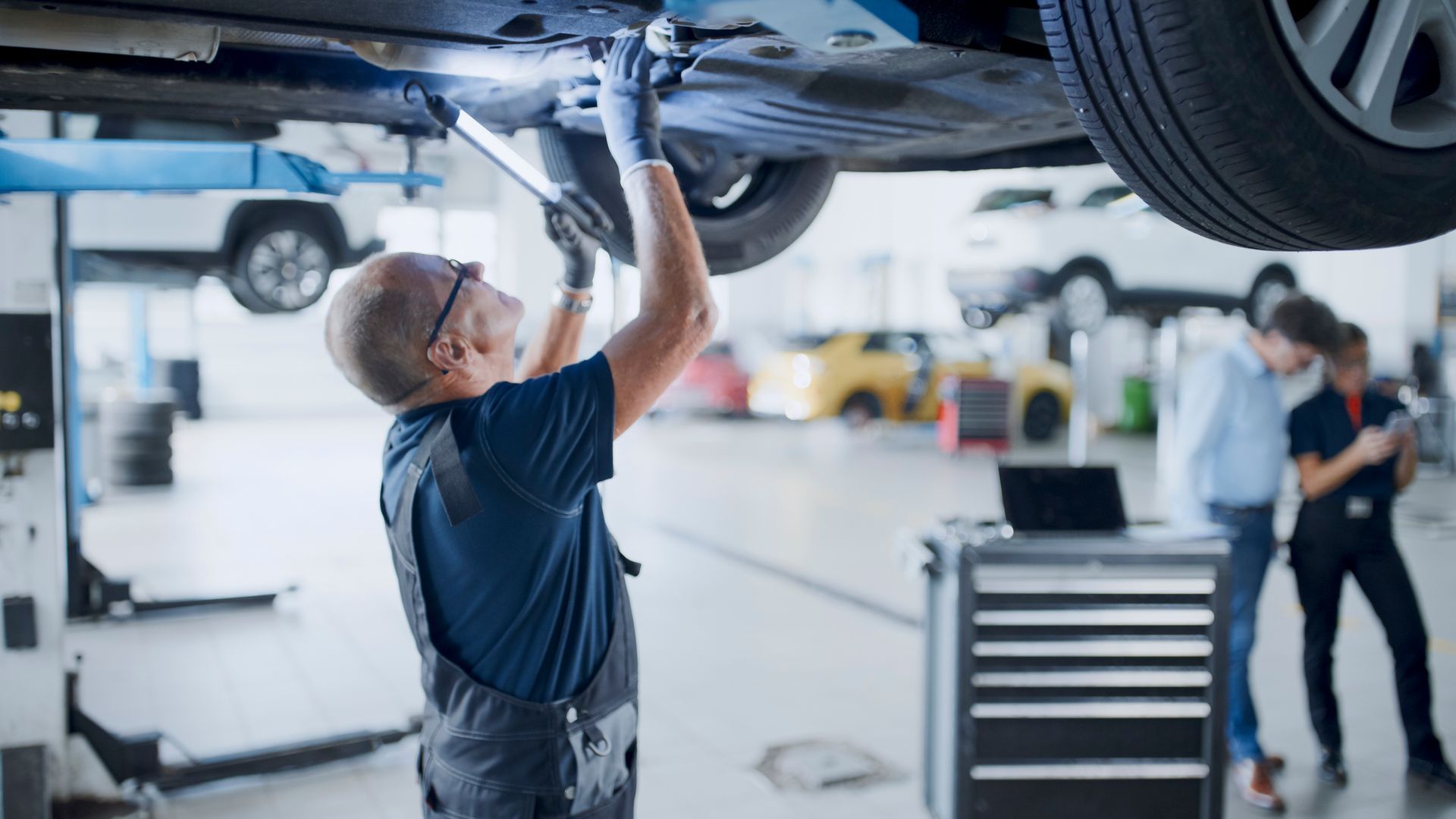 Mechanic working on a car raised on a lift in a garage. Others stand in background.