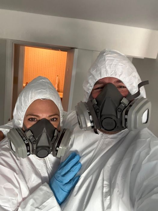 Two people in white hazmat suits and respirators in a room, one with a gloved hand on the other.
